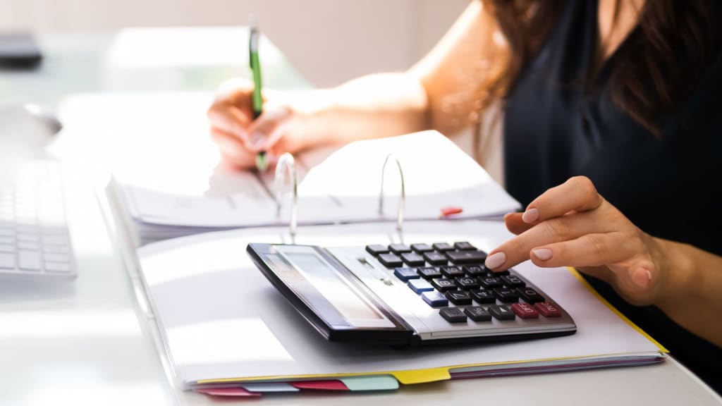 A person uses a calculator and writes on paper with a green pen, seated at a desk with binders.
